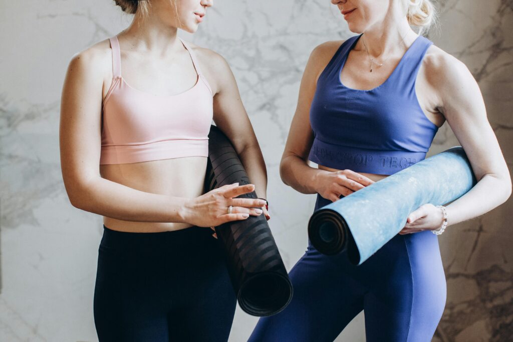 Two women in activewear holding yoga mats, preparing for a yoga session indoors.